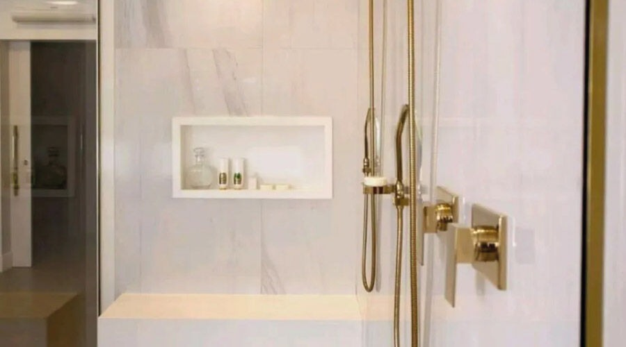 Banheiro com torneiras e detalhas em dourado e nicho fixado na parede contendo shampoo e sabonetes.