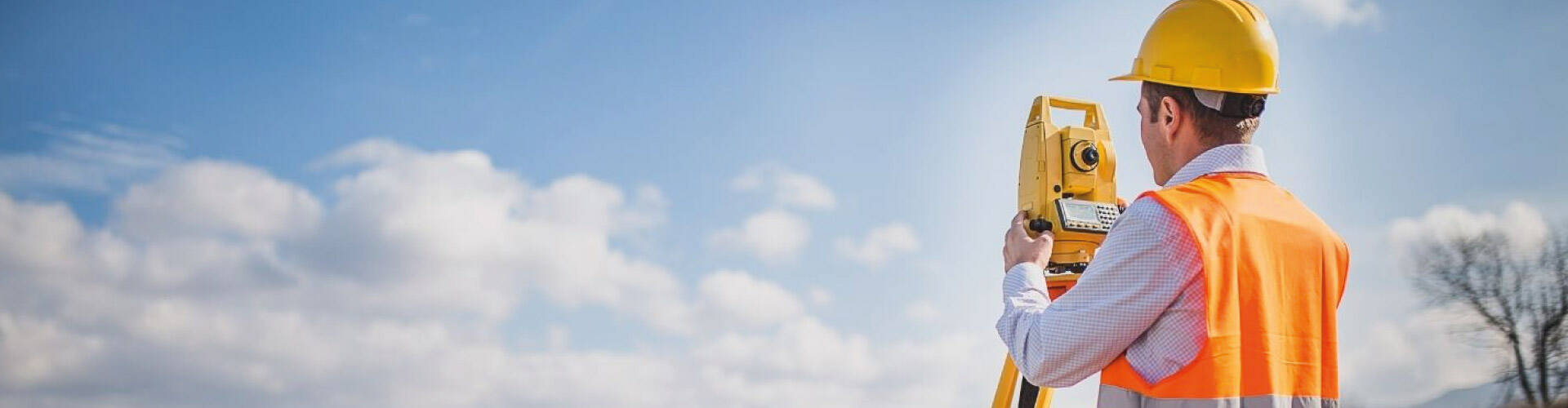 Homem usando colete de segurança laranja em uma estrada, analisando o terreno através do equipamento.