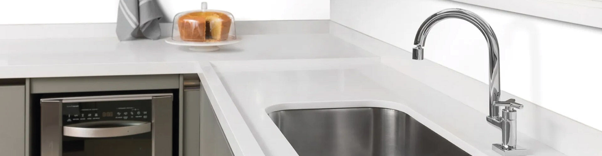 Pia de cozinha moderna com uma torneira de aço inoxidável. Ao fundo, há uma máquina de lava-louças e, em cima do balcão, um bolo em um prato com uma toalha ao lado.